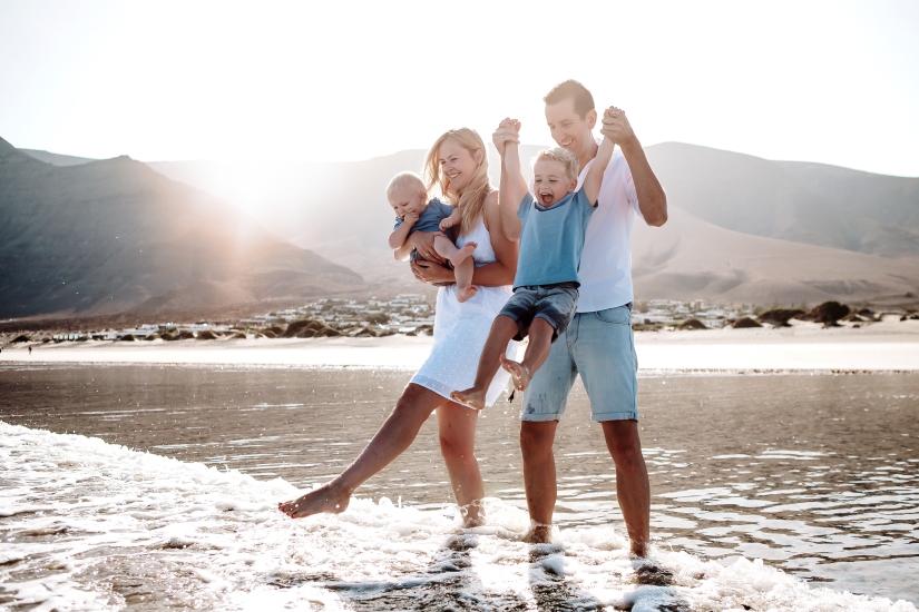 Happy family enjoying the beautiful beach of Famara in Lanzarote, splashing in the water with the sun shining through the mountain in the background. Captured by a professional family photographer in Lanzarote